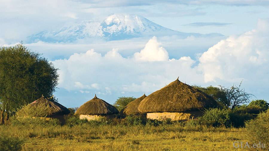 Landscape of huts in a field with the summit of Mount Kilimanjaro emerging from the clouds in the distance, where the December birthstone tanzanite is mined. Landscape of huts in a field with the summit of Mount Kilimanjaro emerging from the clouds in the distance, where the December birthstone tanzanite is mined.
