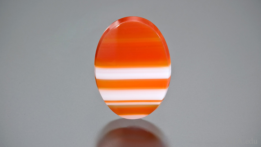 A flat oval polished sardonyx tablet showing the varying orange and white banding of the August birthstone. A flat oval polished sardonyx tablet showing the varying orange and white banding of the August birthstone.